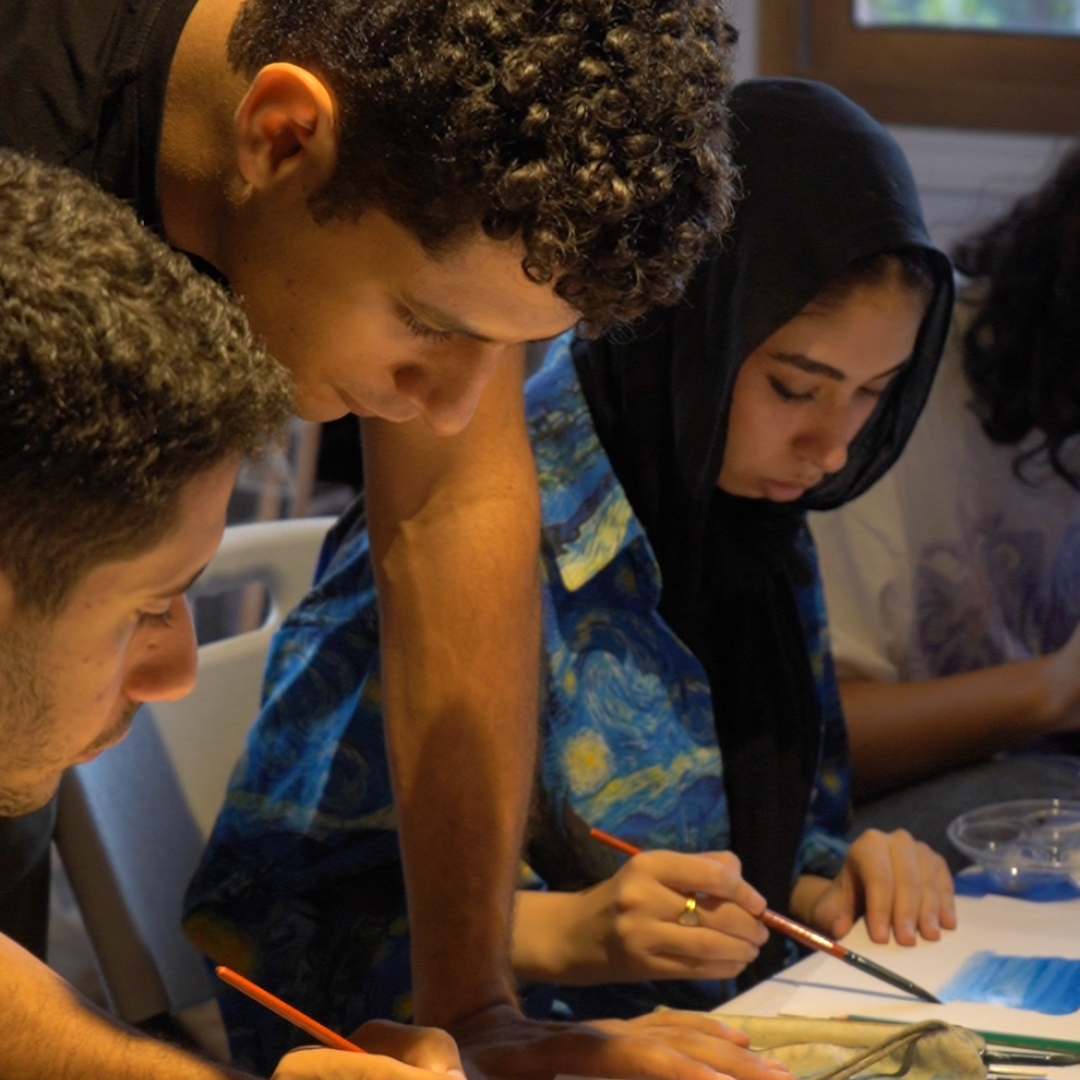 Three people, two men and a woman, are looking at a piece of paper with paintbrushes. One person is wearing a blue and black patterned shirt.